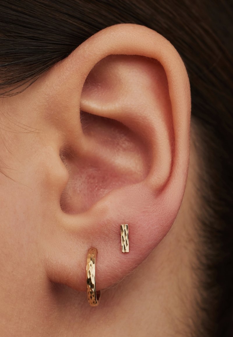Goldene Creolen mit gehämmertem Muster neben einem kleinen, rechteckigen goldenen Stecker. Beide Ohrringe sind glänzend und reflektierend.