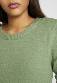 Pull côtelé vert avec un col rond, présentant une texture douce et une coupe ajustée. Vue en gros plan mettant en évidence les détails du tricot.