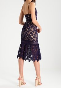 Krajková šaty v námořnické modři s květinovými vzory, tenkými ramínky a rozšířeným lemem. Obsahuje složité detaily a přiléhavý siluetu.