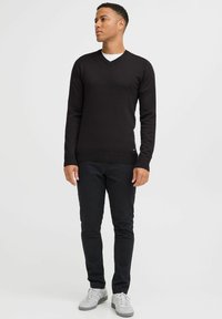 Pulover de culoare neagră cu decolteu în V, cu un design ajustat, confecționat dintr-un material tricotat. Asortat cu pantaloni skinny închiși la culoare și adidași gri, stând în fața unui fundal alb.