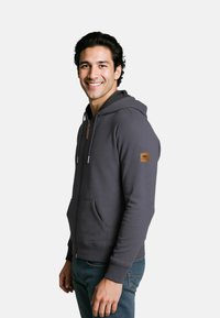 Dark gray zip-up hoodie made from soft fabric, featuring a kangaroo pocket, ribbed cuffs, and a logo patch on the left sleeve.