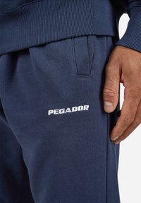 Marineblå joggebukser laget av mykt stoff med ribbestrikket livlinning, med den hvite broderte logoen "PEGADOR" på siden.