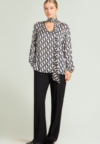 Langærmet bluse med bølget sort og creme mønster, V-hals, påsyet tørklædedetalje; kombineret med sorte skræddersyede bukser og sorte sko.