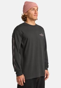 Langärmliges schwarzes T-Shirt aus Baumwolle, mit pinkem Logo auf der Brust und gemusterten Akzenten entlang der Ärmel; kombiniert mit einer gerippten pinken Mütze.
