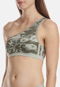Sostén deportivo verde de un solo hombro con patrón de camuflaje texturizado, banda elástica y detalle de logo en la parte frontal, diseñado para ropa activa.