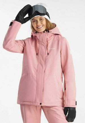 Smilende person i lyserød vinterjakke og bukser, hvid hue, løfter sorte skibriller med handske mod hvid baggrund.