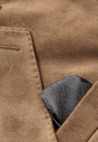 Barna velúr anyag, varrt zsebbel, amelyben egy hajtogatott fekete mintás zsebkendő látható, kiemelve a textúrát és a színkontrasztot.