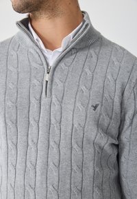 Maglione grigio a maglia con colletto con zip, caratterizzato da motivi a treccia e dettagli a costine sottili. Piccolo logo visibile sul petto.