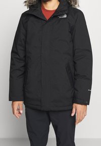 Veste d'hiver noire avec une capuche en fourrure synthétique, fermeture éclair, poches latérales et poignets élastiques ; présente un logo sur le haut de la poitrine et sur la manche.