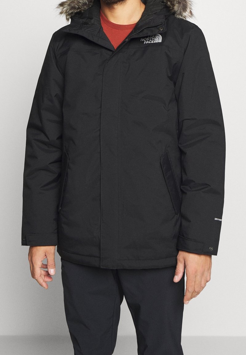 Veste d'hiver noire avec une capuche en fourrure synthétique, fermeture éclair, poches latérales et poignets élastiques ; présente un logo sur le haut de la poitrine et sur la manche.