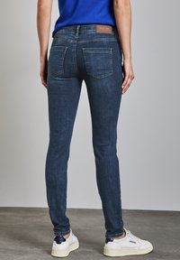 Jean skinny bleu foncé avec une texture lisse, comportant deux poches arrière et une étiquette en cuir marron sur la taille.