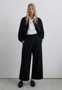 Black oversized jacket with two front pockets, white shirt, and high-waisted black pleated trousers. Black loafers complete the outfit.