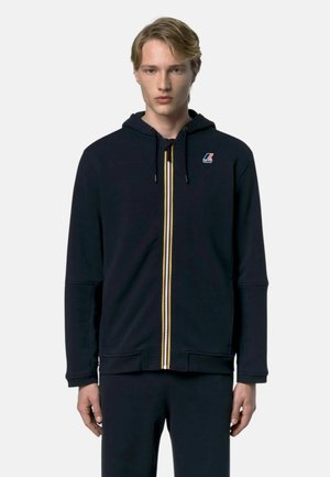 Giovane uomo che indossa una felpa nera con cappuccio e zip verticale tricolore con un piccolo logo sul petto, in piedi su uno sfondo semplice.