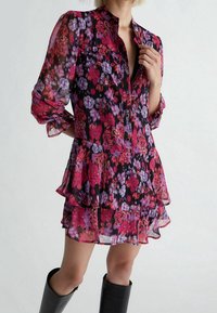 Vestito camicia floreale in tessuto trasparente con tonalità di rosa, viola e nero. Presenta una chiusura con bottoni, colletto e volant sovrapposti all'orlo.