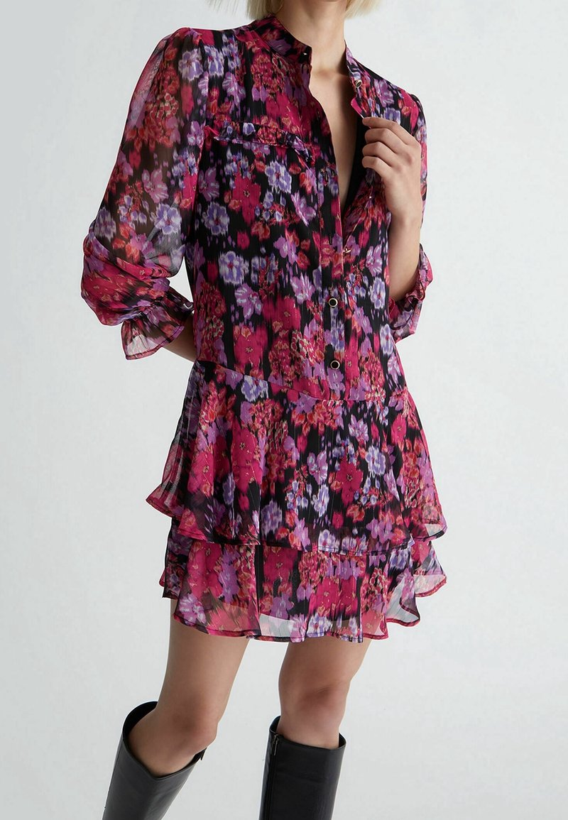Vestito camicia floreale in tessuto trasparente con tonalità di rosa, viola e nero. Presenta una chiusura con bottoni, colletto e volant sovrapposti all'orlo.