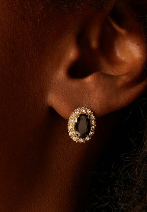 Close-up van een donkerhuidig oor met een gouden oorbel met een grote zwarte centrale steen omringd door kleine heldere kristallen.