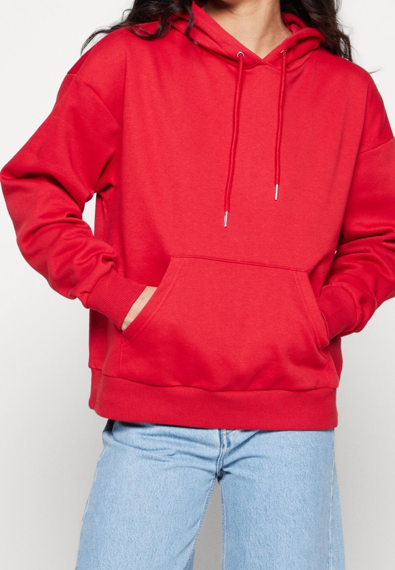 Femme portant un sweat à capuche rouge vif avec poche avant et un jean bleu clair, les mains dans les poches du sweat.