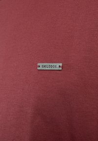 Metalen label met het opschrift "SMILODOX" bevestigd aan een gestructureerde, bordeauxrode stof achtergrond. Het label heeft een eenvoudig rechthoekig ontwerp met insnijdingen.