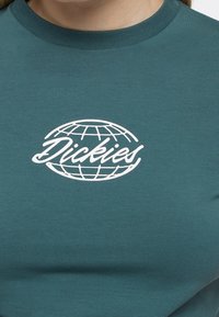 Tyrkysové bavlněné tričko s klasickým výstřihem. Na přední straně se nachází bílý design globu se slovem "Dickies" napsaným kurzívou uprostřed.
