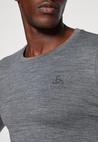 Camiseta cinza de gola redonda feita de um tecido texturizado. Logótipo discreto impresso no peito. Vista em close que foca no material e na gola.