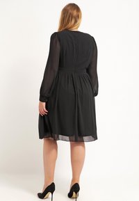 Czarna sukienka z długimi, przezroczystymi rękawami, marszczonym pasem i spódnicą do kolan. Gładka tekstura i detal z zamkiem błyskawicznym z tyłu. Noszona z czarnymi szpilkami.