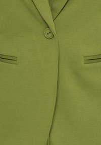 Un seul bouton vert fixé sur un blazer vert clair avec des revers crantés et deux poches passepoilées étroites à l'avant.
