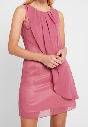 Bezrękawna mini sukienka w kolorze przytłumionego różu z warstwowymi, plisowanymi zdobieniami z przodu, noszona przez osobę z rękami splecionymi na talii.