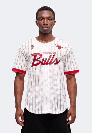 Homme portant un maillot de baseball blanc des Chicago Bulls avec des fines rayures rouges et des logos, debout devant un fond clair uni.