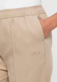 Beige bukser med en glat tekstur, elastisk talje, sidelommer og et diskret broderet logo nær lommen.