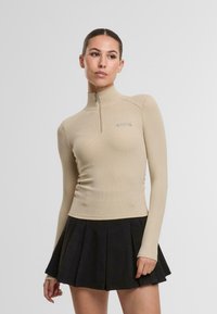 Šviesiai rudas ilgomis rankovėmis zip-up viršus su suraityta tekstūra, turintis aukštą apykaklę ir prigludusį dizainą, derinamas su juoda klostuota mini sijonu.