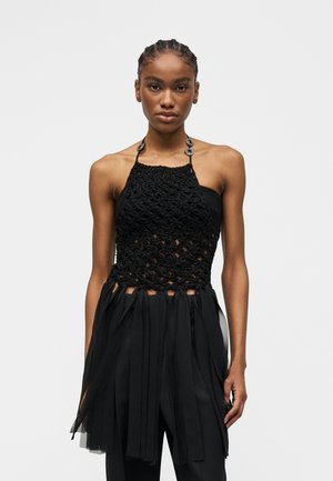 Kobieta z zaplecionymi włosami, ubrana w czarny, bezrękawnikowy top z dekoltem halter, o fakturowanym, siateczkowym wzorze i warstwowymi frędzlami.