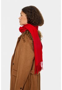 Kvinde med krøllet hår iført en brun frakke og lys rød tørklæde, vendt til venstre på en ensfarvet hvid baggrund.