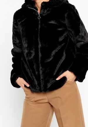 Person wearing a black faux fur zip-up jacket with hood and camel-colored pants, hands in jacket pockets.