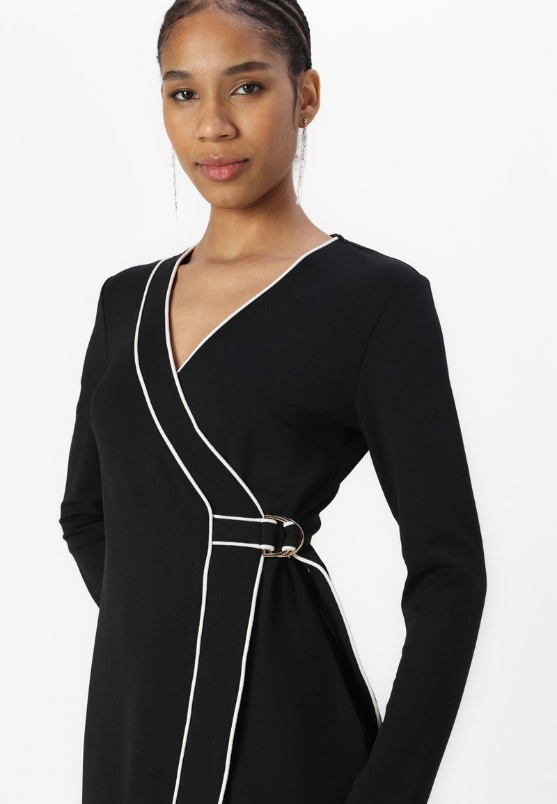 Vestido negro ajustado con ribetes blancos, mangas largas y un detalle de hebilla lateral. Tela suave con un diseño elegante y entallado.