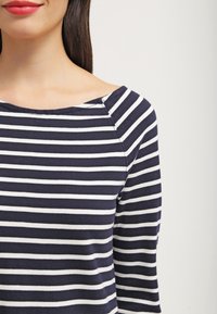 Femme aux longs cheveux foncés portant un haut à manches longues à rayures horizontales bleu marine et blanc, col bateau, sur un fond clair uni.