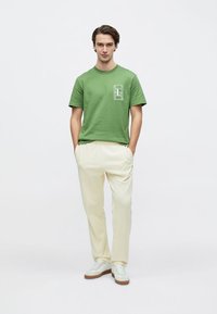 Πράσινο βαμβακερό μπλουζάκι με κοντά μανίκια και λευκό γραφικό σχέδιο στο στήθος, συνδυασμένο με μπεζ παντελόνι και λευκά αθλητικά παπούτσια.