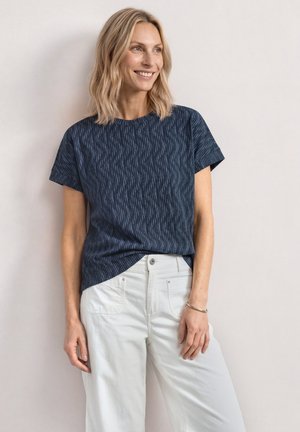 Femme blonde souriante, portant une chemise à manches courtes bleu foncé à motifs, rentrée dans un pantalon blanc taille haute, debout contre un mur clair.