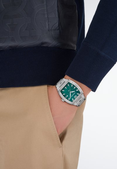 Tengerészkék pulóver texturált bőrpaneleként, bordázott mandzsettákkal; ezüst karóra türkiz számlappal, dátumfunkcióval és masszív szíjjal; bézs nadrág.