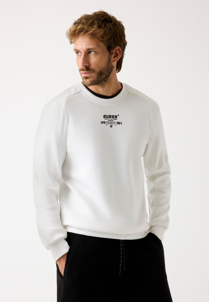 Balts džemperis ar teksturētu audumu, apaļu kakla izgriezumu un melnu "GUESS" logo ar 1991. gadu. Modelis valkā melnas bikses ar kabatām.