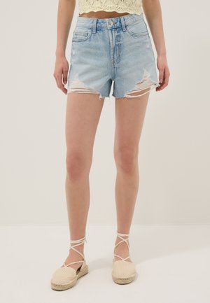 Femme portant un short en jean délavé bleu clair, un crop top en crochet crème et des espadrilles beiges avec liens à la cheville, debout sur un fond uni.