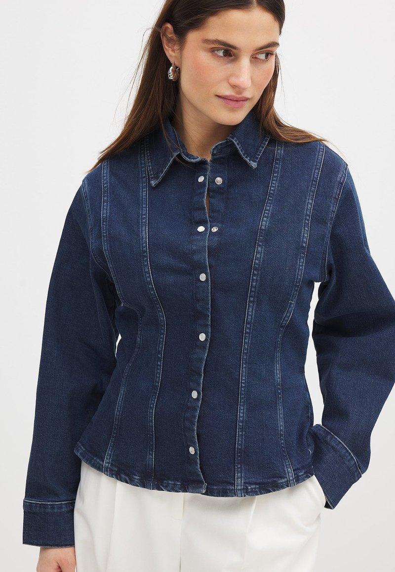 Dark denim button-up shirt featuring a structured design, pointed collar, and contrasting stitch lines. Silver buttons down the front.