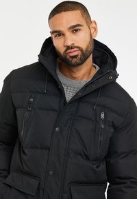 Chaqueta acolchada negra hecha de tejido suave, con cierre de cremallera, capucha ajustable y dos bolsillos frontales. Presenta un diseño acolchado.