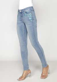 Jeans ajustados de color azul claro con adornos de lentejuelas a lo largo de los bolsillos. Textura de denim suave, cintura de altura media y diseño estándar de cinco bolsillos.