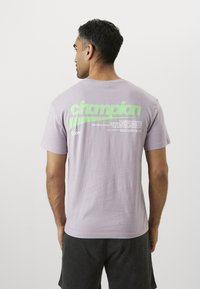 Lavendel bomull t-skjorte med en stor grønn "champion" logo på baksiden, med ekstra tekst og grafikk nedenfor.