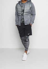 Grå zip-up pufferjacka, grå kamouflagemönstrad långärmad tröja, svarta shorts, kamouflagemönstrade leggings och vita träningsskor.