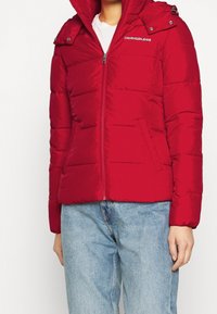Rote Steppjacke mit Reißverschluss und Seitentaschen. Verfügt über eine Kapuze und das Calvin Klein Jeans Logo auf der Brust. Glatte Textur, gestepptes Muster.
