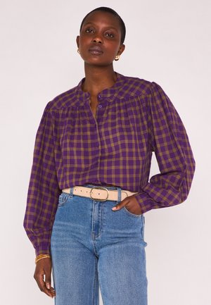 Vrouw draagt een paars geruit overhemd met lange mouwen, gestoken in een blauwe spijkerbroek met een beige riem, staand met één hand in de zak.