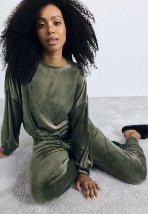 Olive green, velvety loungewear set with long sleeves and pants, featuring a relaxed fit. Model sitting on a soft, light-colored surface.