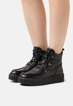 Sorte læderankelstøvler med ribbede, tykke såler; har snørebånd og metalbeslag som pynt. Overfladen har en glat tekstur.