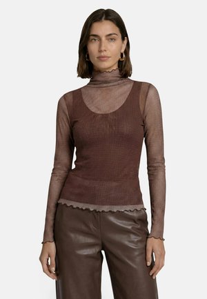 Femme portant un haut marron en maille à manches longues avec un col roulé et un ourlet ondulé, ainsi qu'un pantalon en cuir marron, debout, les bras détendus.